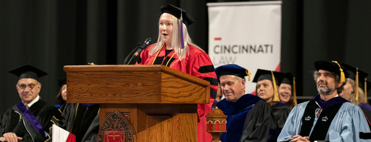 a speaker stands behind a podium during a graduation ceremony, with a seated group of people behind them, all wearing graduation gowns. 